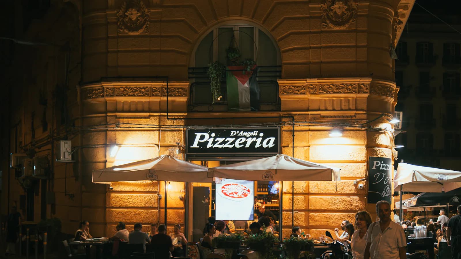 Pizzeria napolitaine illuminée de nuit, façade et terrasse extérieure, couple qui marche à proximité